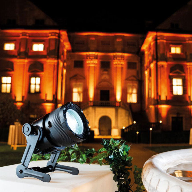 Astera QuikSpot LED uplight in outdoor event setting illuminating building facade with warm orange light mounted on QuikTurn yoke bracket on table with greenery and table setting at night